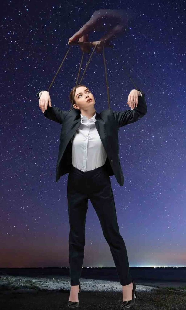 A human woman, standing, with strings attached to her like a marionette puppet. She is looking up and can barely see the faint image of the hand that controls her strings.