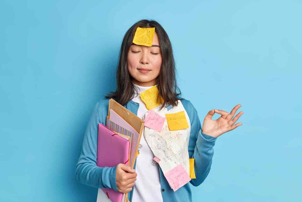 A woman standing with her eyes closed. Her right arm is full of files and a notebook computer. Her left hand is raised to about her shoulder. Her left hand is doing the meditation sign: her ring finger is touching her thumb, and her remaining fingers are out straight. She also has post-it notes all over her front, even one on her forehead…but she is calm.