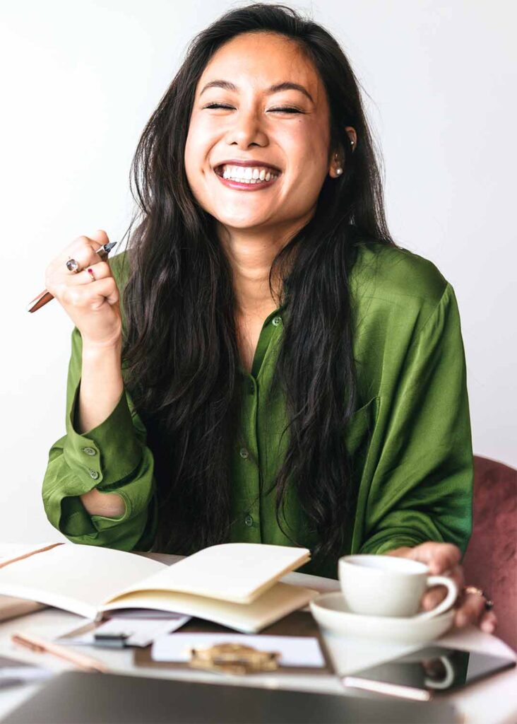 A smiling woman sitting at a desk with a pen in hand and a blank notebook in front of her. She is happy because she is about to use The To-Do List Secret.